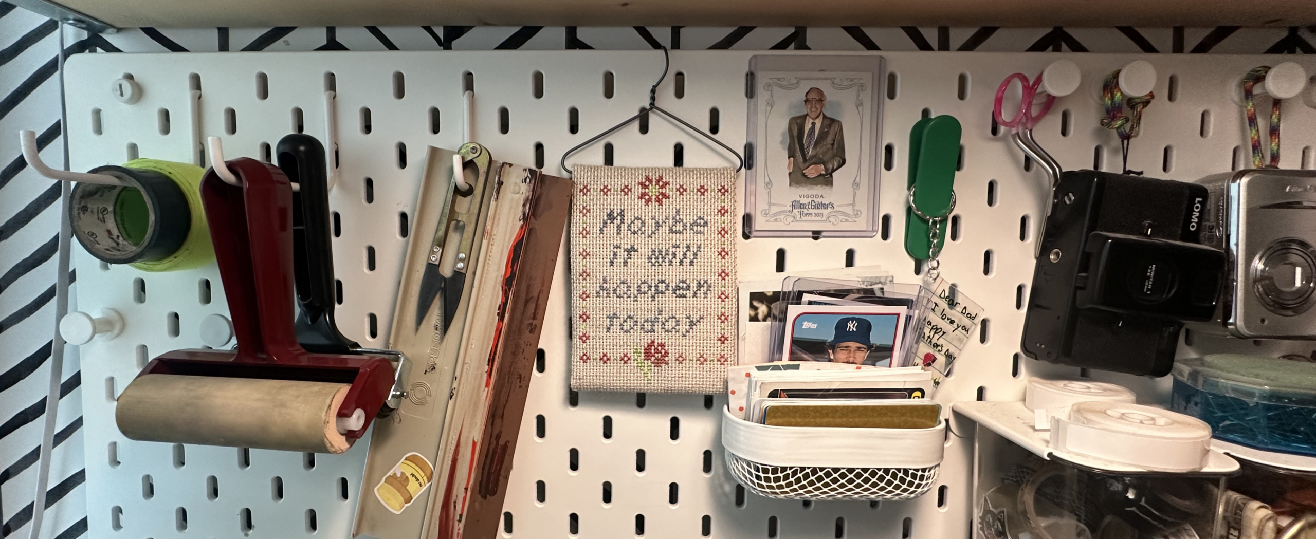 A photo of a pegboard with various tools and supplies, and in the middle a finished cross-stitch on a hanger that reads "Maybe it will happen today" surrounded by a floral border.